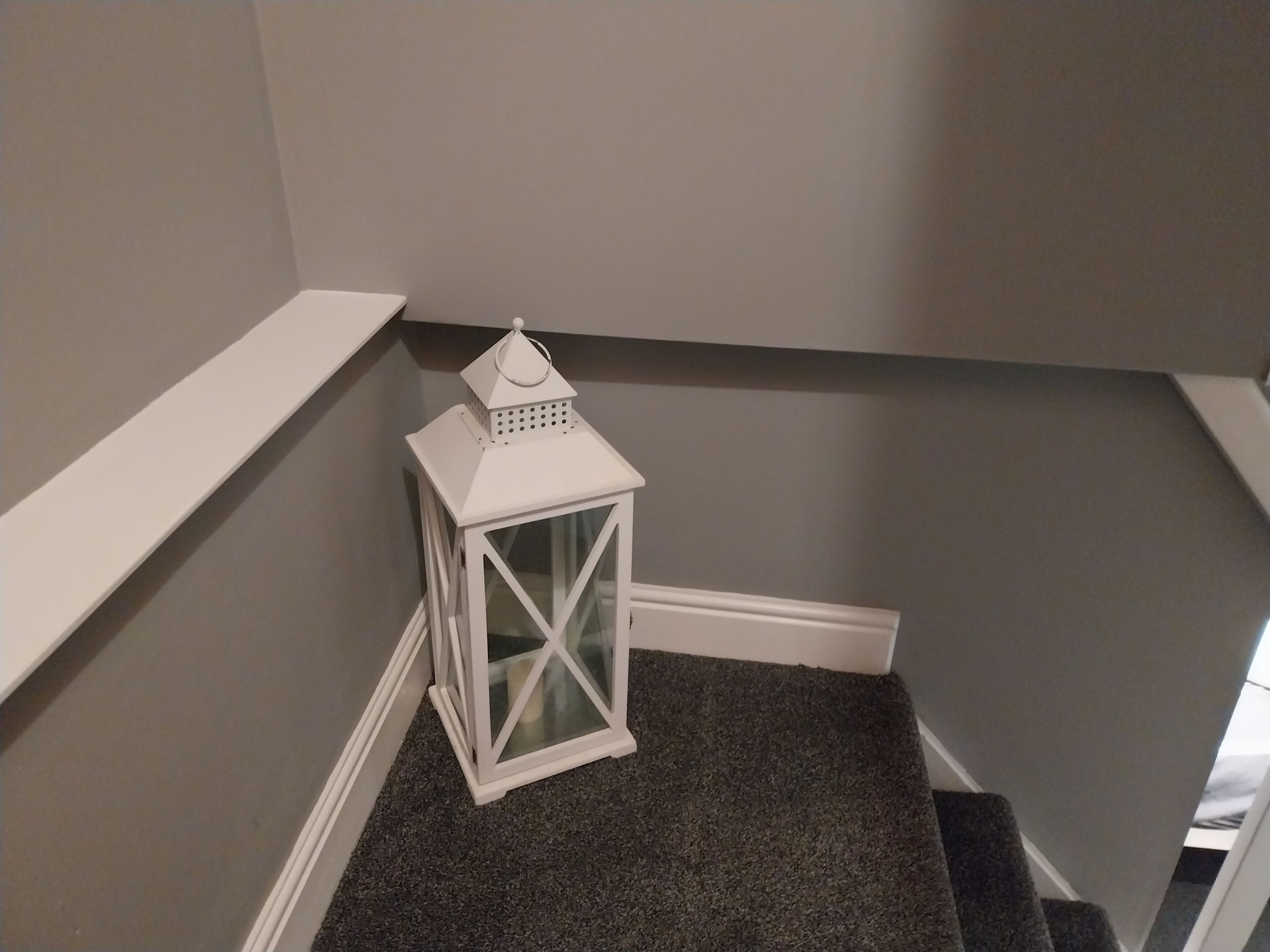 Eine weiße Laterne mit Glaskuppel auf einem dunklen Teppichboden in einer Zimmer Ecke, mit weißen Wänden und einer weißen Fußleiste.