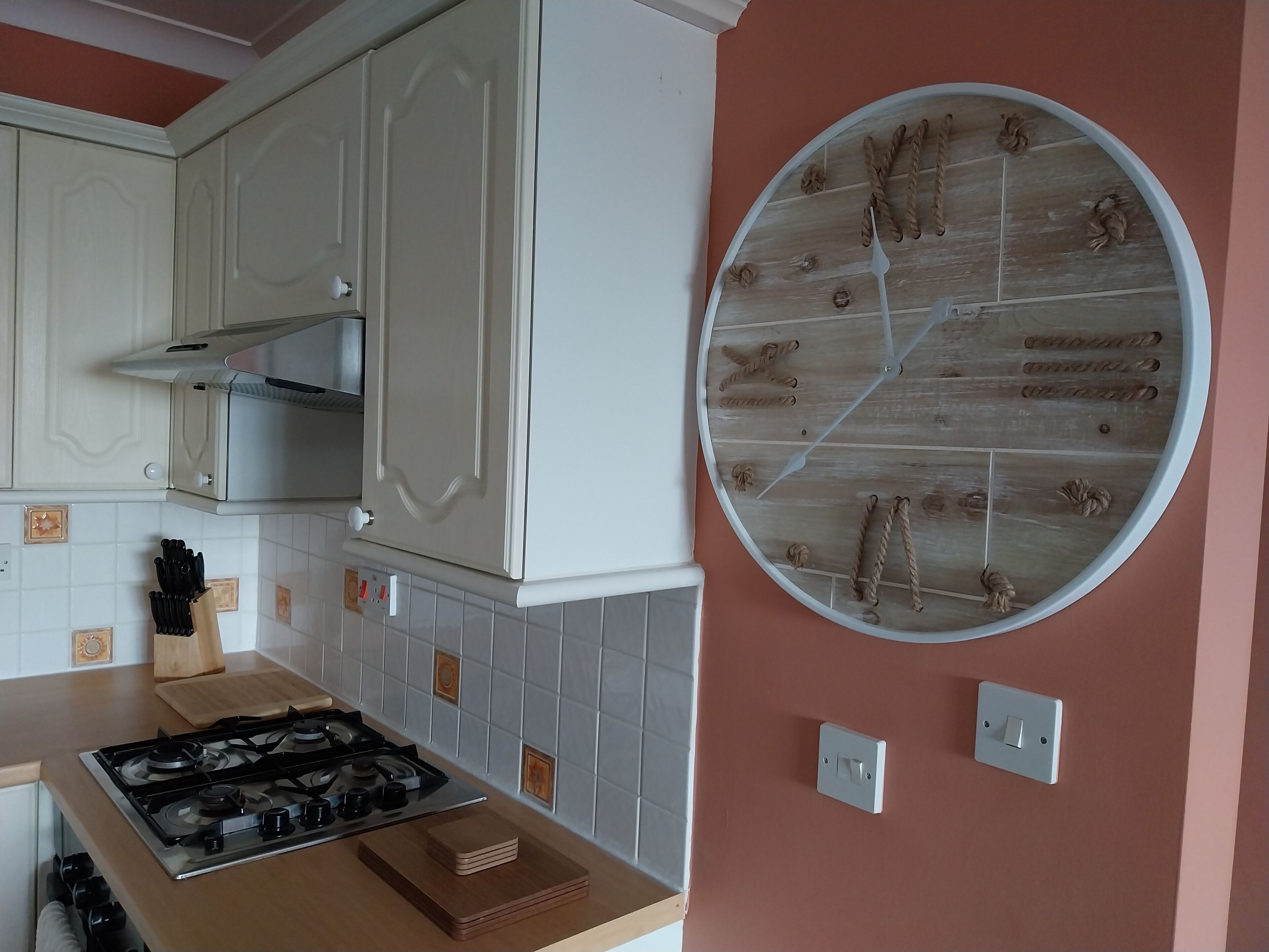 Una cocina con armarios blancos, estufa a gas, tabla de cortar de madera y un reloj redondo sujeto a la pared.