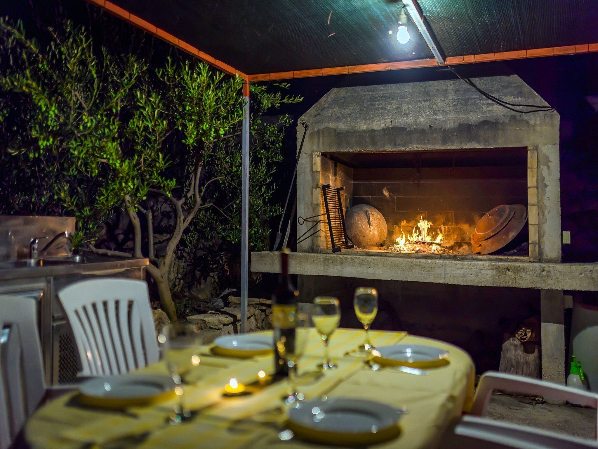 An outdoor dining area with a stone oven, a table set for a meal, and a backdrop of greenery.