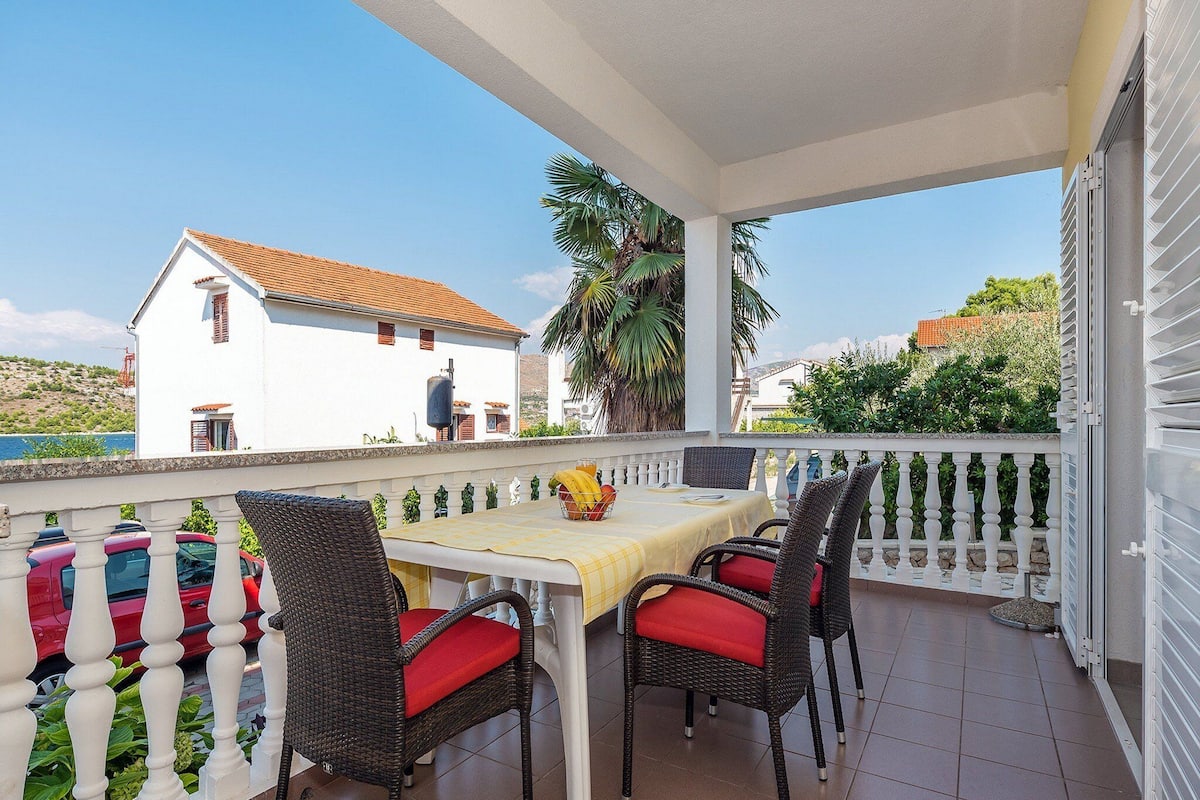 A balcony with a table and four wicker chairs, a white house in the background, and a clear view of the sea.