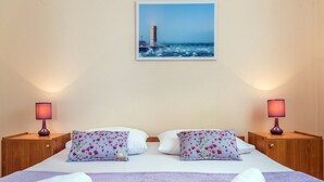 A hotel room with a bed, two nightstands, two lamps, two pillows, a folded towel, and a painting of a lighthouse on the wall. - Apartments Gulin (Sibenik)