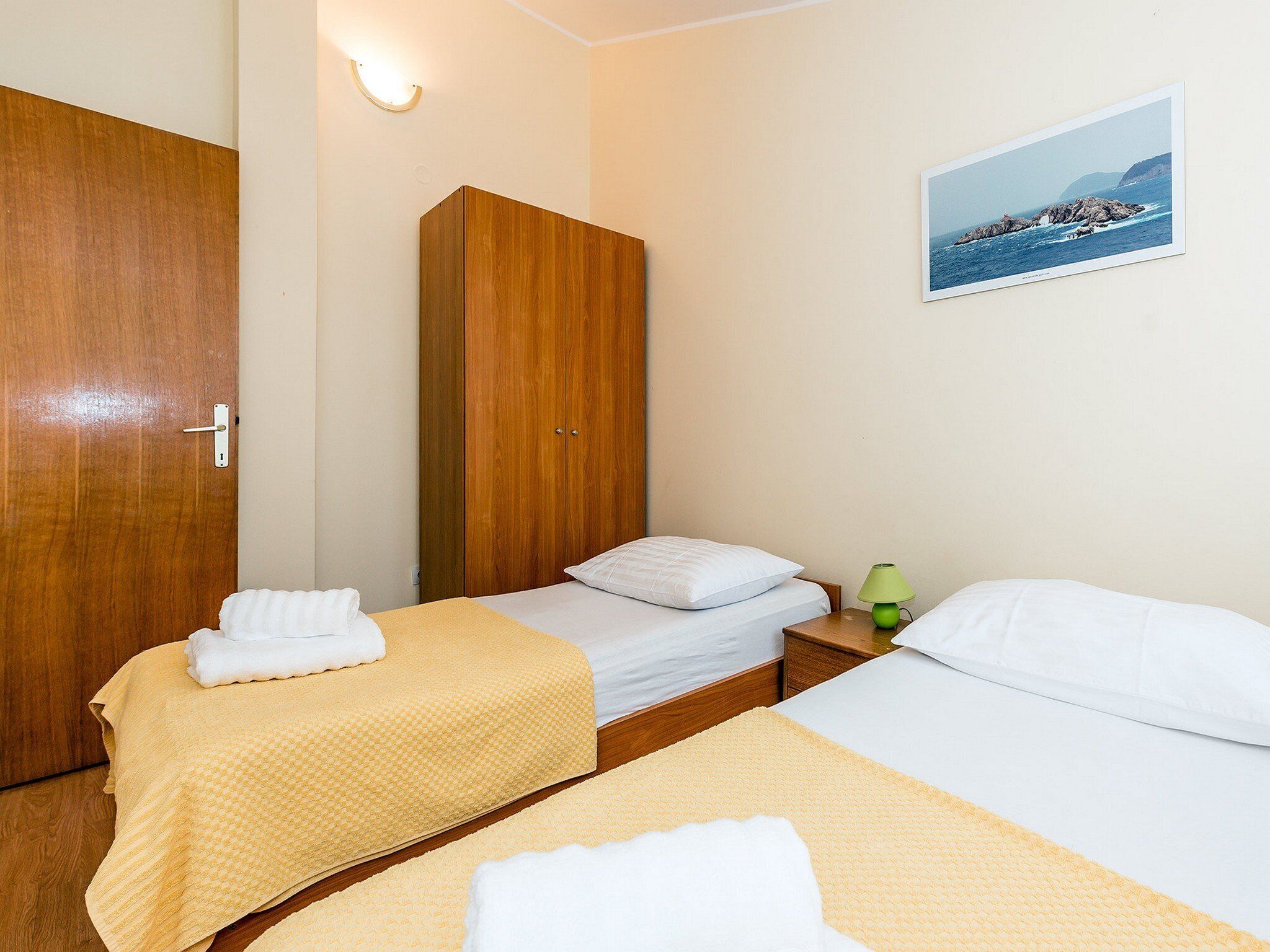 Una habitación de hotel con dos camas individuales, un armario de madera, una mesita de noche con lámpara y un cuadro en la pared.