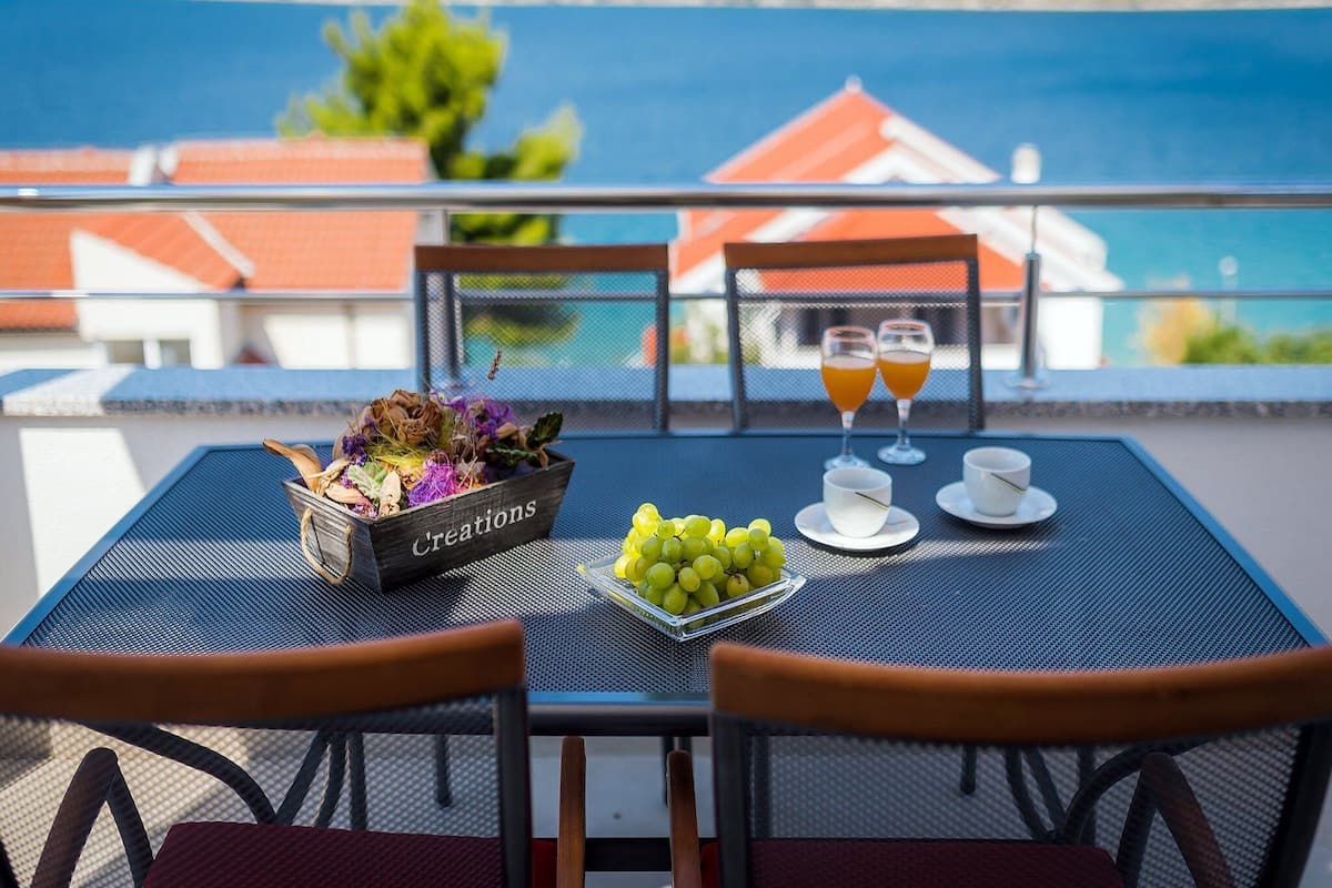 A table with a bowl of grapes, two glasses of orange juice, and a tray of grapes on a balcony overlooking a body of water and buildings.