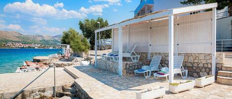 Une terrasse en bord de mer avec un sol en pierre, des fauteuils blancs et un espace salon couvert, le tout adossé à un mur en pierre.