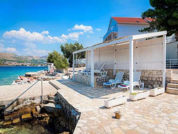 A coastal terrace with stone flooring, white lounge chairs, and a covered seating area with a stone wall backdrop. - Apartments Gulin (Sibenik)