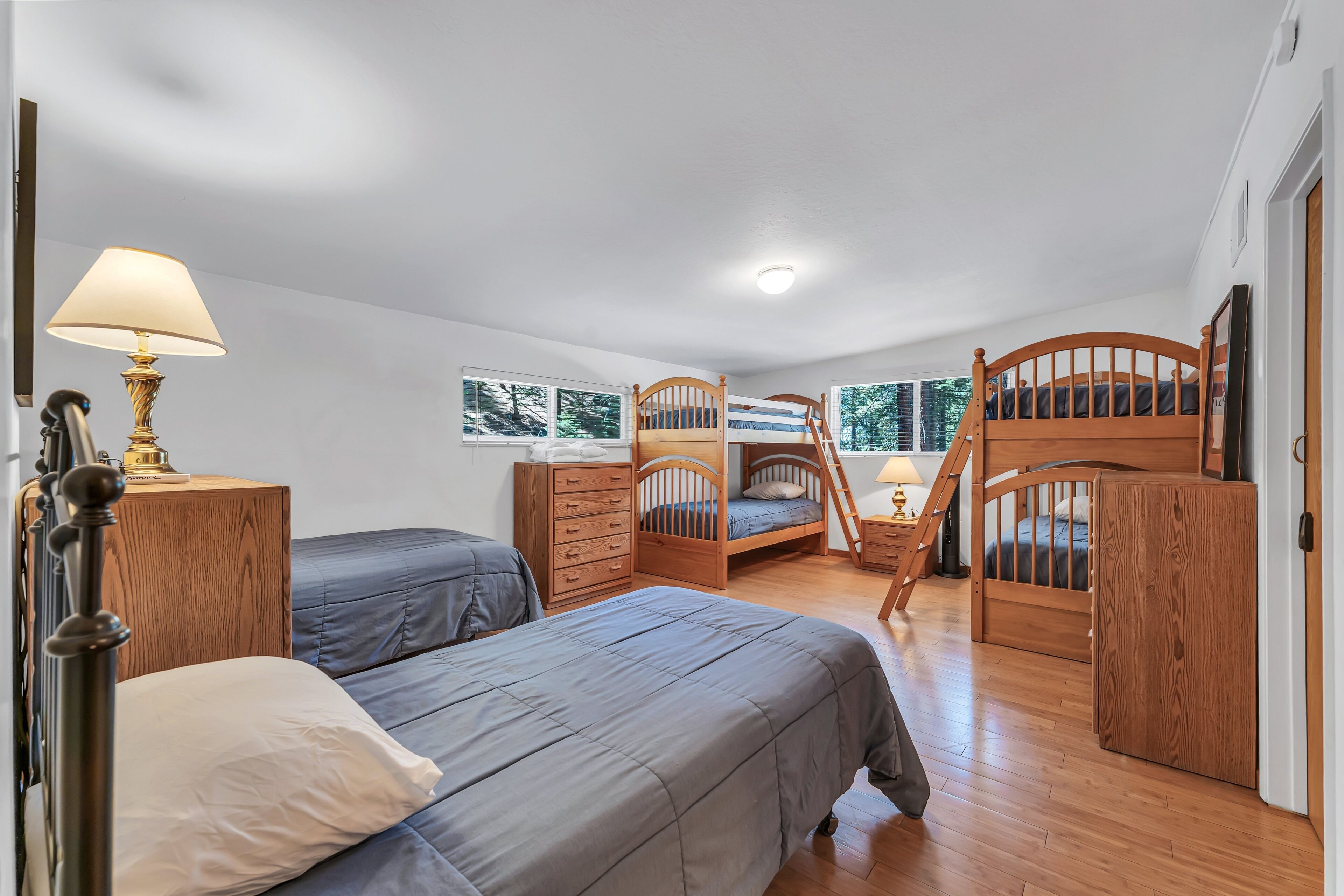 A bedroom with two beds, a wooden dresser, a nightstand, a lamp, and a window with a view of trees.