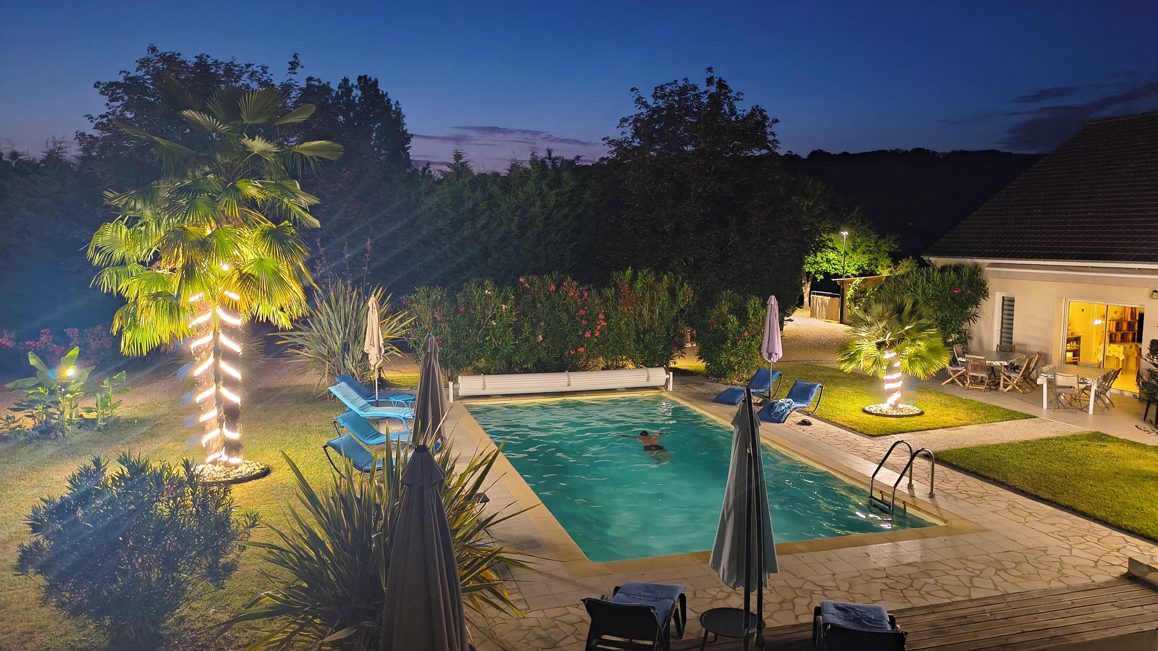 A swimming pool with lounge chairs and umbrellas, surrounded by greenery and a building with a tiled roof.