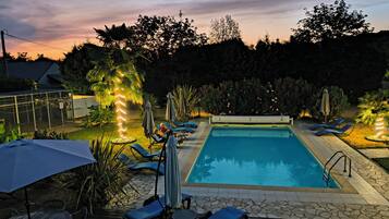 A swimming pool with lounge chairs and umbrellas, surrounded by greenery and a sunset sky.