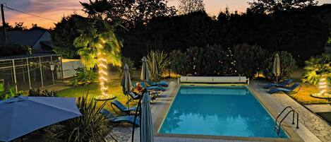 A swimming pool with lounge chairs and umbrellas, surrounded by greenery and a sunset sky.