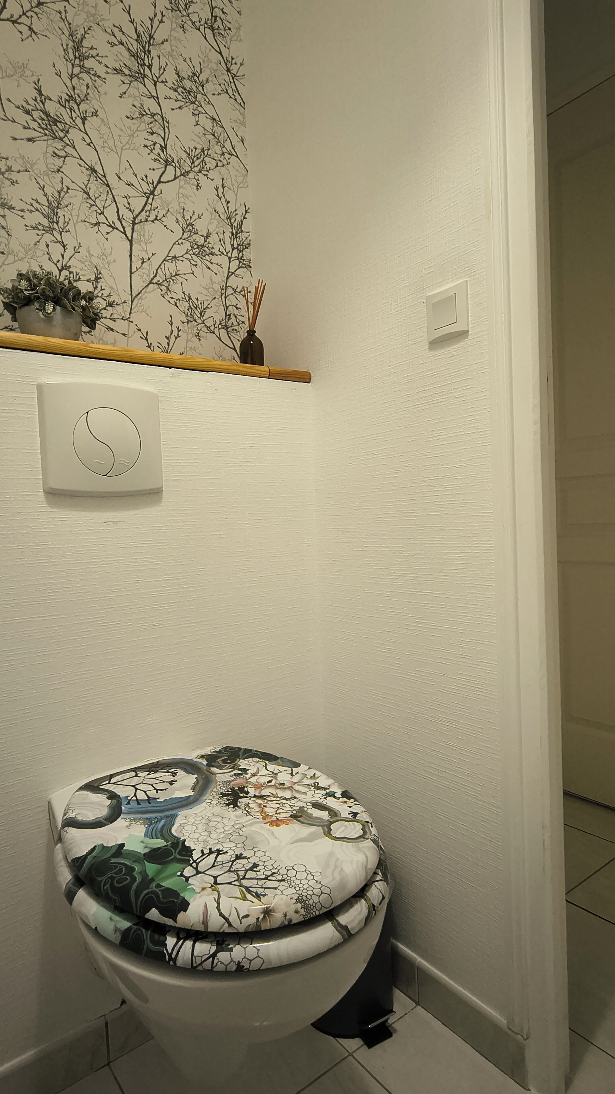 Une salle de bain comprenant des toilettes, un interrupteur fixé au mur et une étagère décorative agrémentée d’une plante et d’un diffuseur de parfum à bâtonnets.