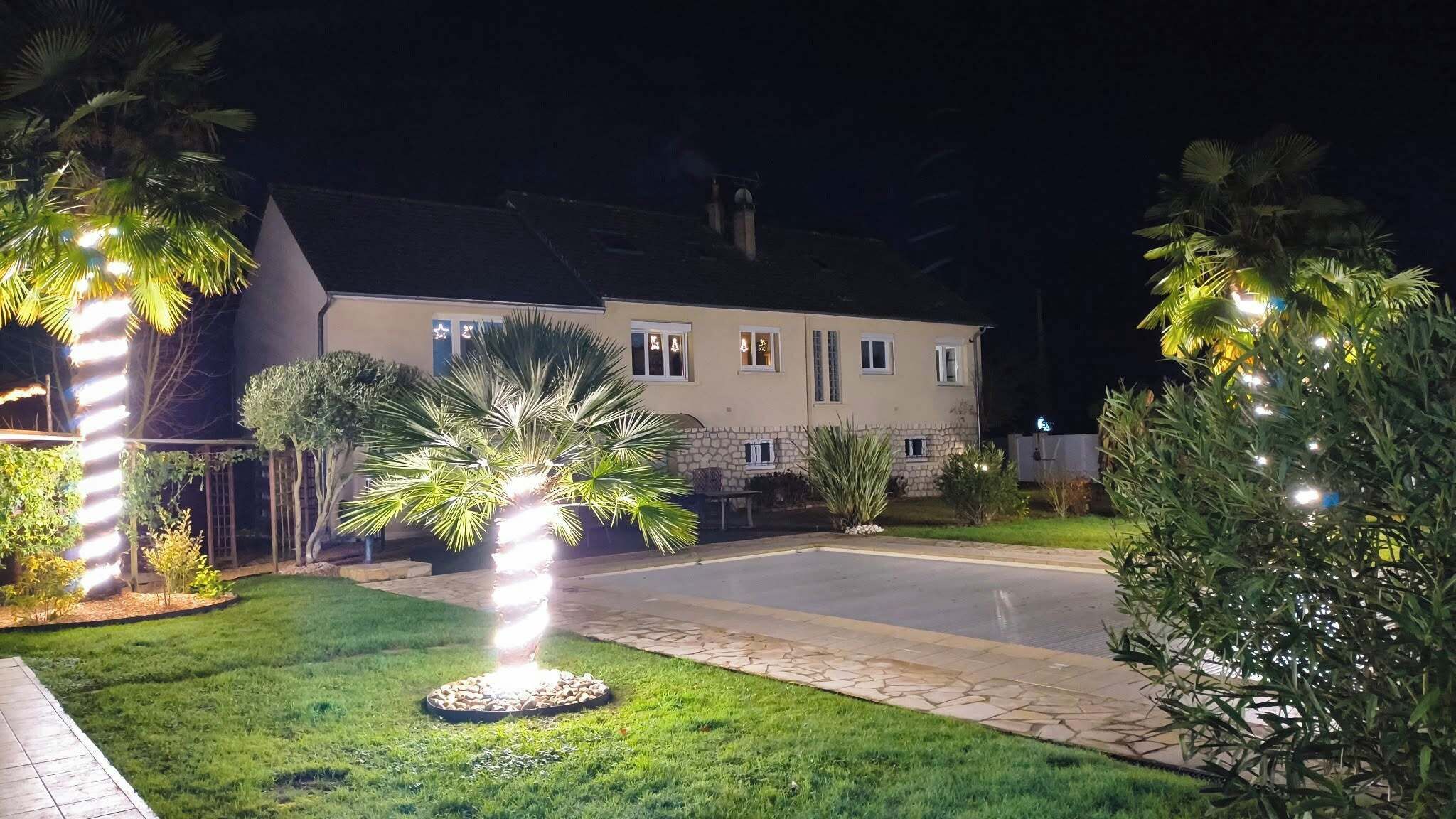 A well-lit residential property at night with a paved driveway, landscaped garden, and a house with multiple windows and a stone facade.