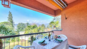 Un balcón con una mesa dispuesta para dos personas, sillas blancas y vistas a zonas verdes y montañas.