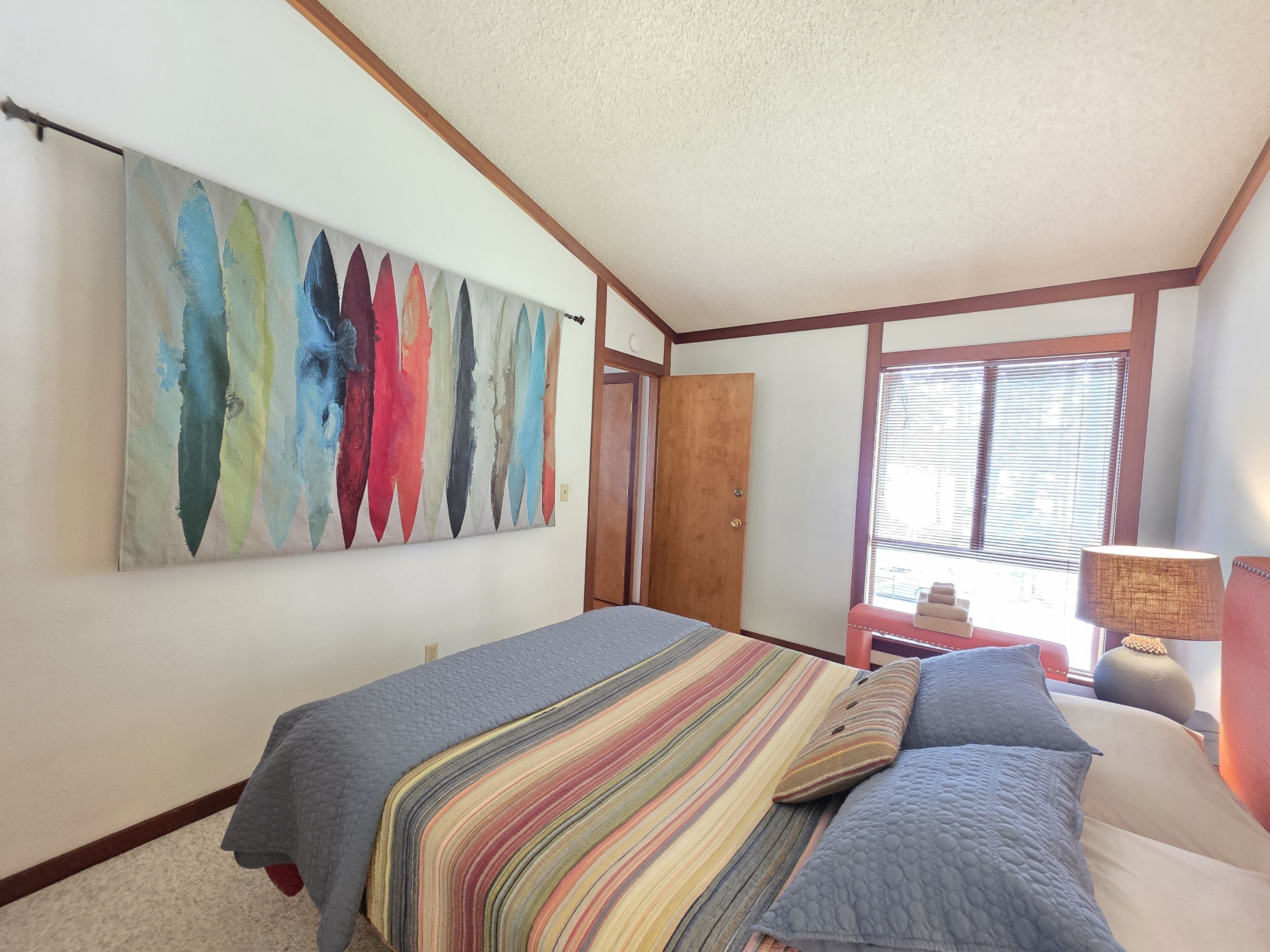 A bedroom with a bed, a colorful abstract painting, a window with blinds, and a nightstand with a lamp.
