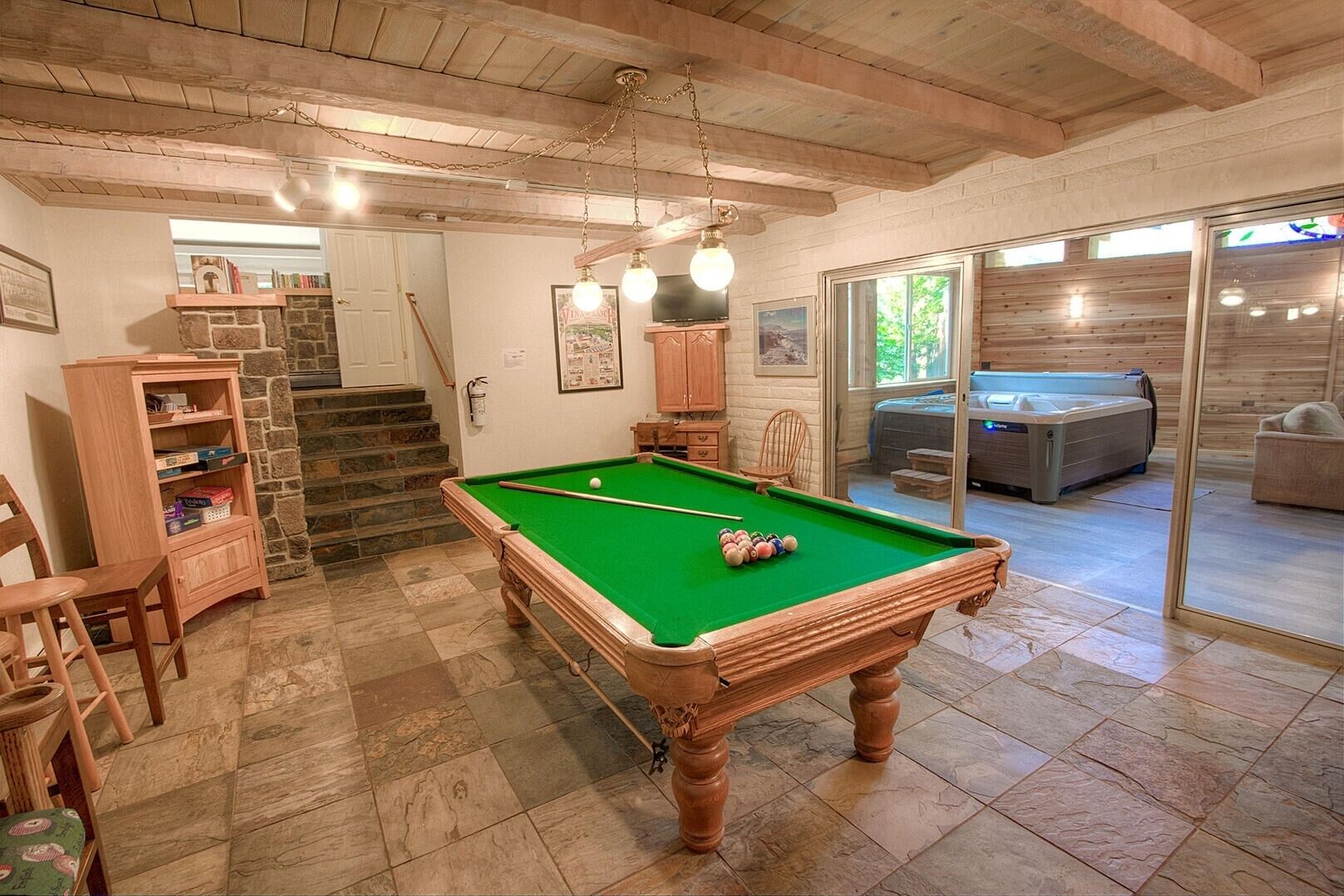 Une table de billard dans une pièce aux murs en pierre, au plafond orné de poutres en bois et à laquelle un escalier mène vers un autre espace.