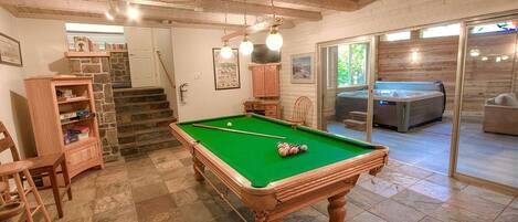 Une table de billard dans une pièce aux murs en pierre, au plafond orné de poutres en bois et à laquelle un escalier mène vers un autre espace.