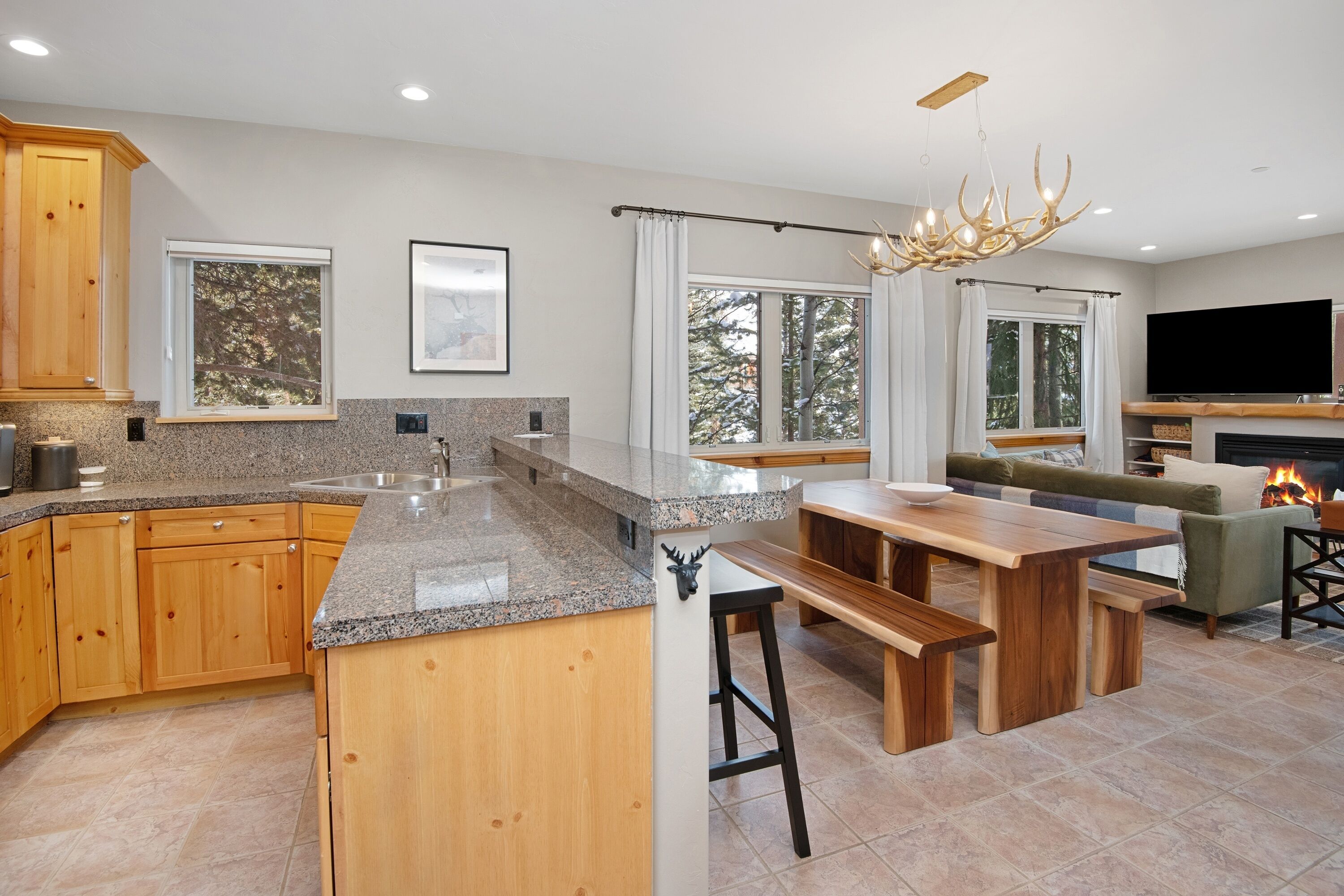 A modern kitchen with granite countertops, wooden cabinets, and a central island. There is a dining area with a wooden table and benches, and a fireplace with a TV above it.