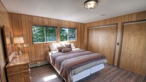 A bedroom with a wooden paneled wall, a bed with a striped bedspread, two windows, a nightstand with a lamp, and a wardrobe.