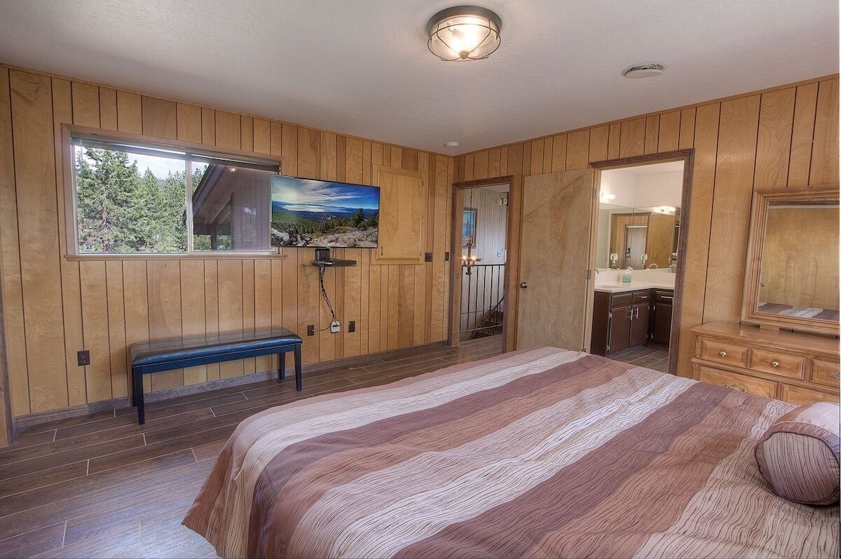A bedroom with a large bed, wooden walls, a bench, a painting, and a bathroom visible through an open door.