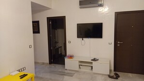Une pièce avec un téléviseur fixé au mur, une console blanche, une table jaune avec des télécommandes et une porte.