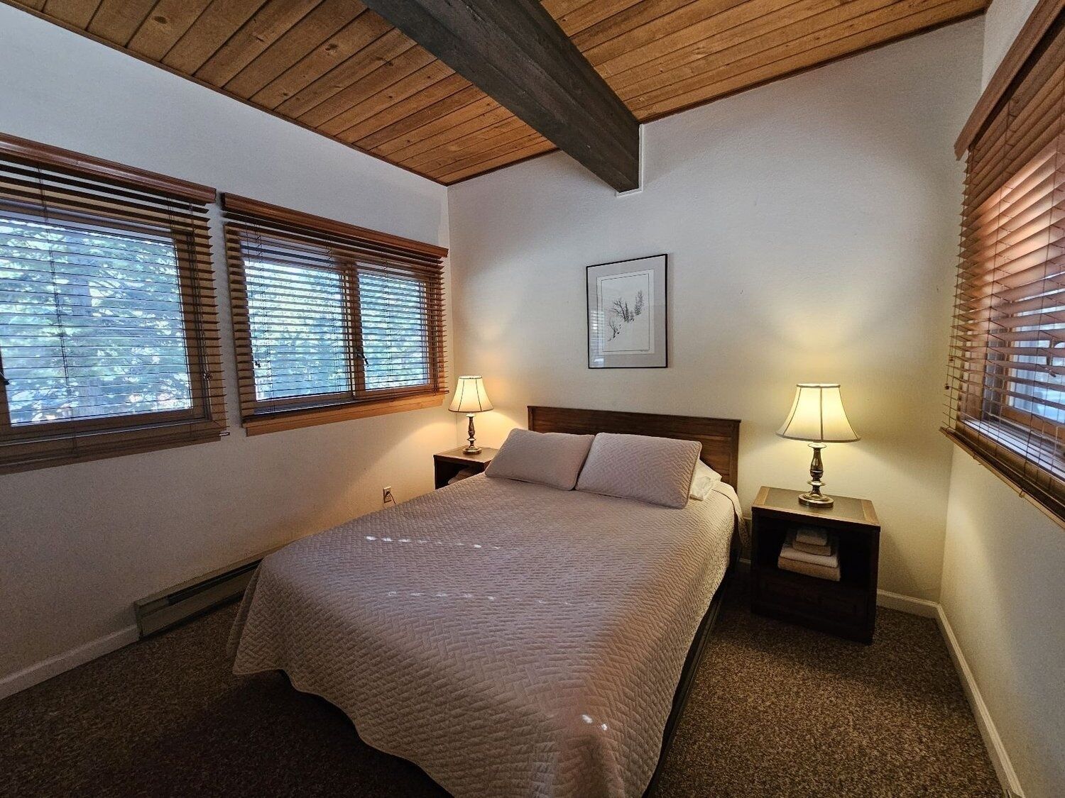 A bedroom with a bed, two bedside tables with lamps, wooden ceiling beams, and windows with blinds.