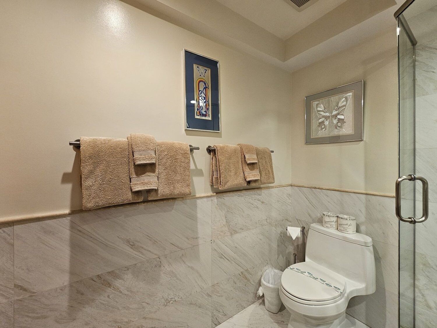 A bathroom with a toilet, two towels, a framed picture, and a glass shower door.
