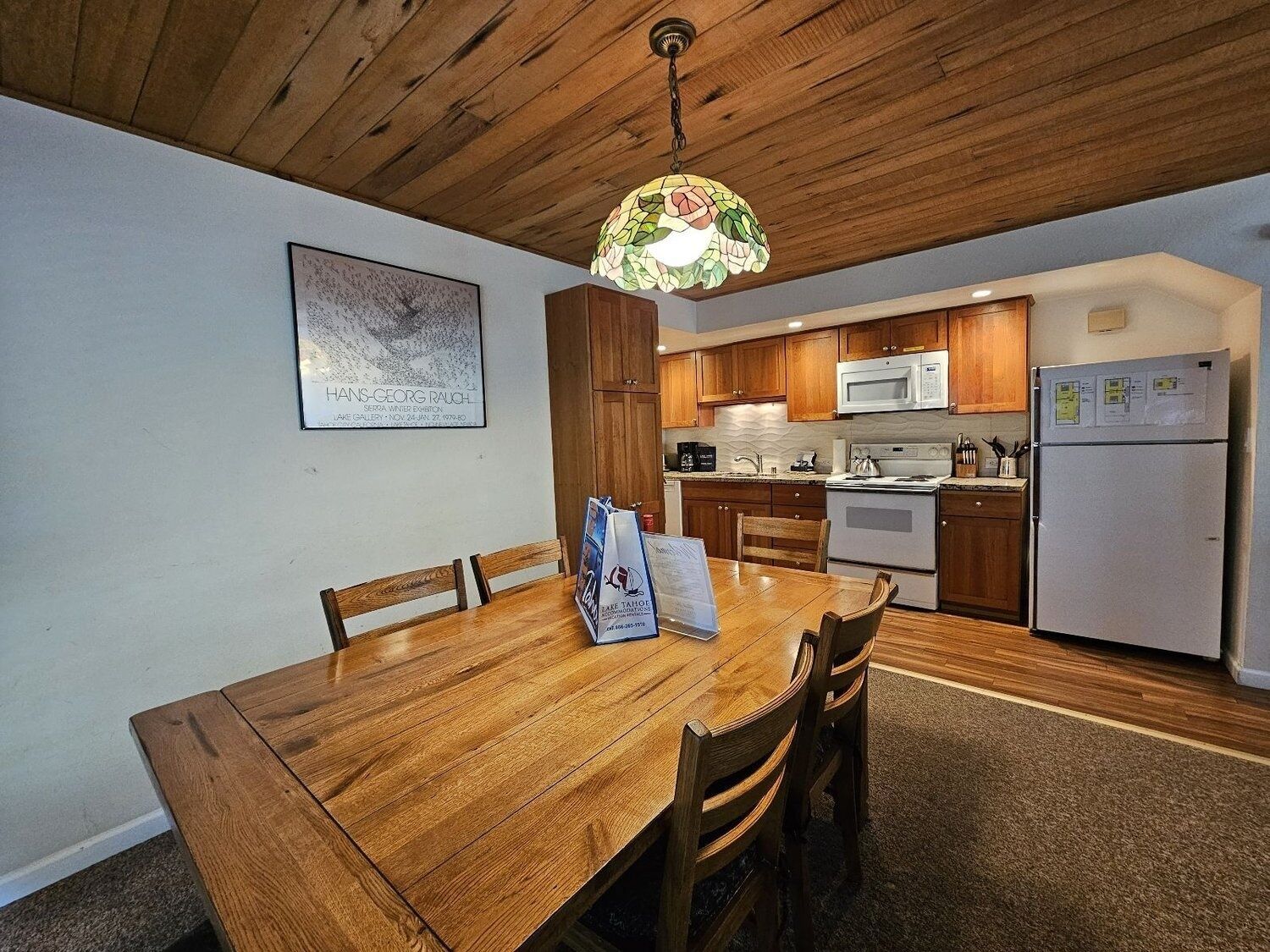A wooden dining table with chairs, a kitchen with wooden cabinets, a microwave, and a refrigerator.