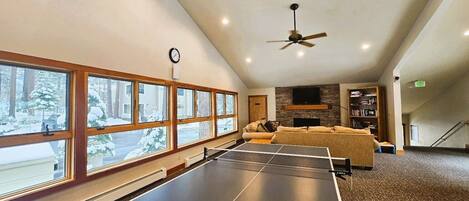 Une table de ping-pong dans une pièce dotée de grandes fenêtres, d’un ventilateur de plafond et d’une cheminée.