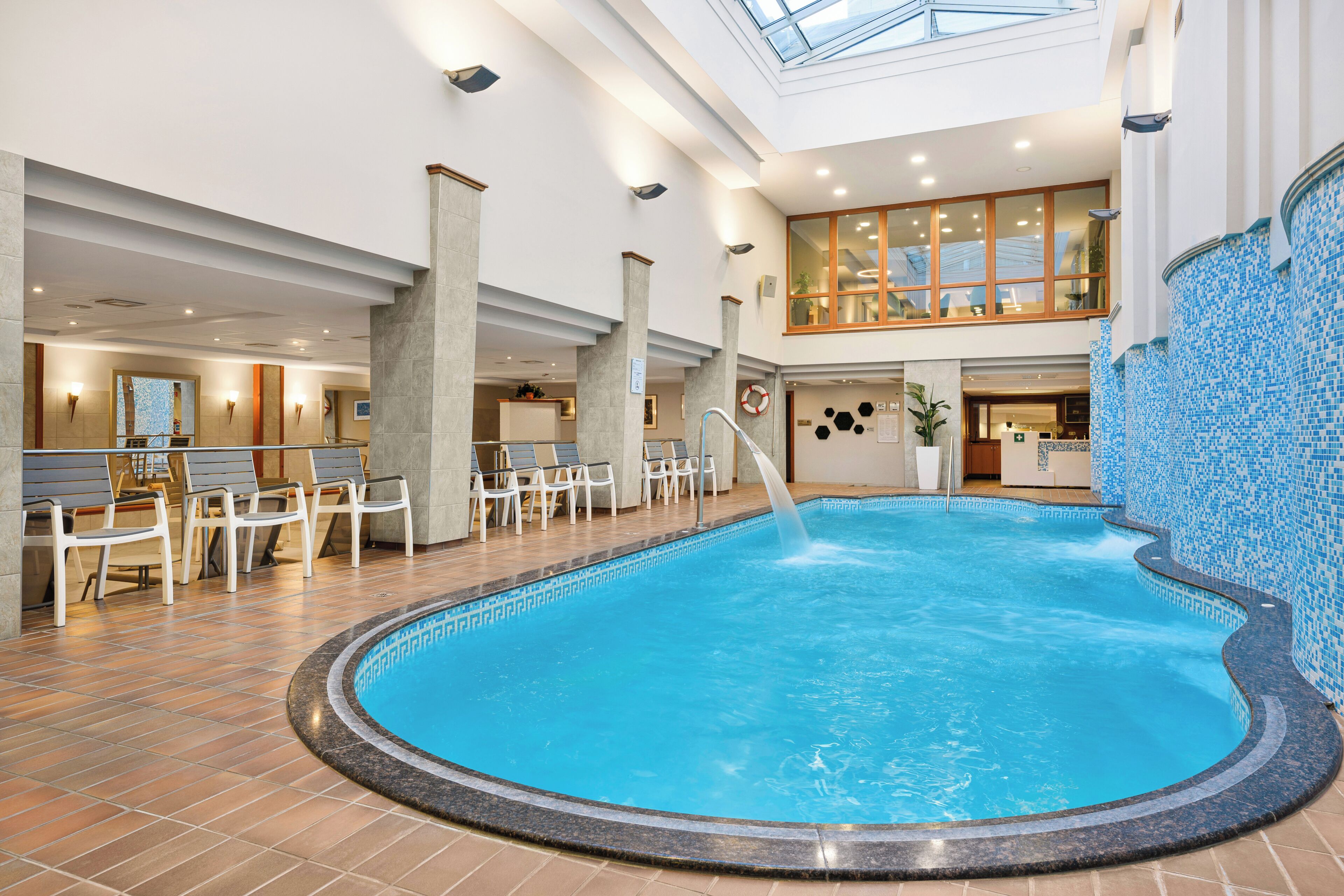 An indoor swimming pool with a tiled floor, lounge chairs, and a high ceiling with skylights.