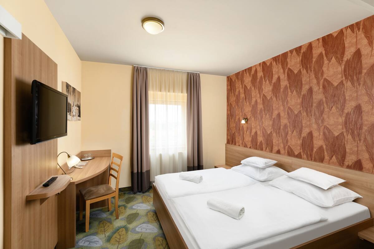 Habitación de hotel con cama, escritorio, silla, televisor y ventana con cortinas.