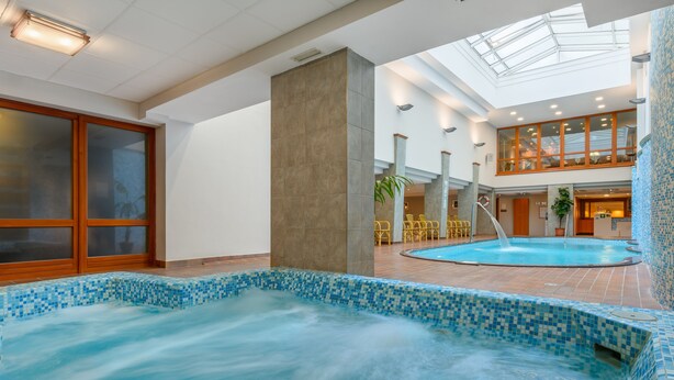 A large indoor swimming pool with a tiled edge, a high ceiling with skylights, and a seating area with yellow chairs. - Aranyhomok Hotel Business & Relax (Kecskemet)