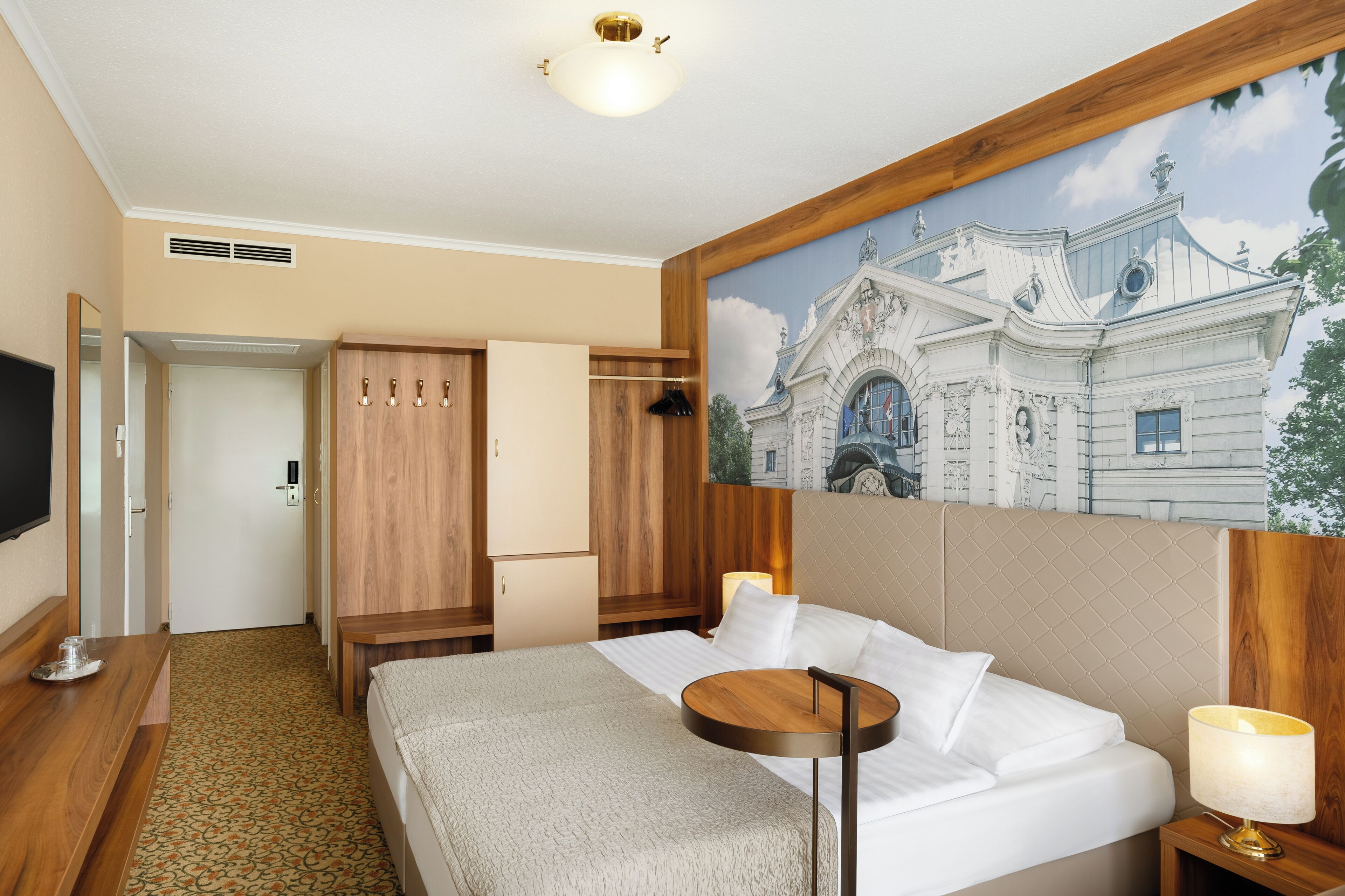 A hotel room with a bed, a wooden wardrobe, a desk with a lamp, and a painting of a building on the wall.