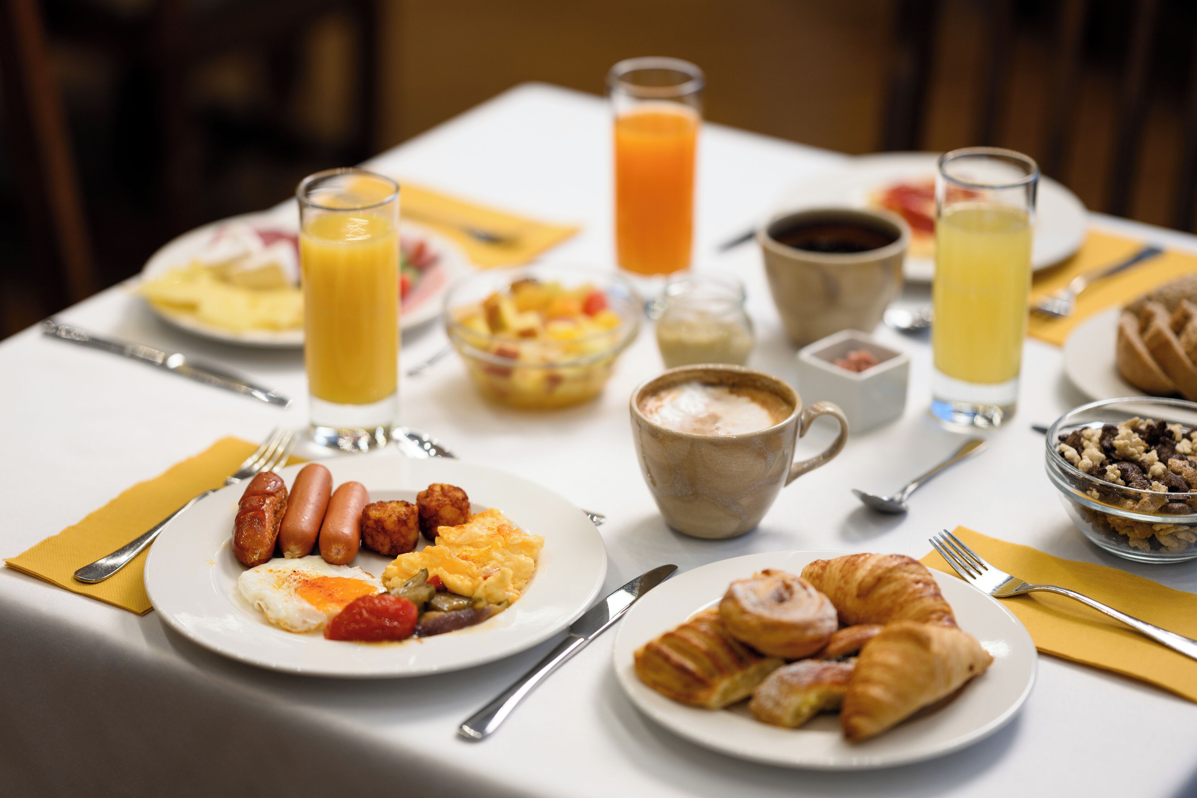 A breakfast table with plates of food, including sausages, eggs, and croissants, along with glasses of orange juice and coffee.