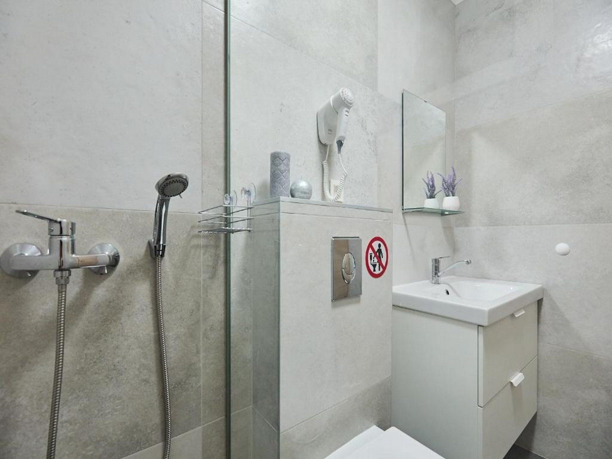 A modern bathroom with a glass shower enclosure, a handheld showerhead, a hairdryer, and a sink with a mirror above it.