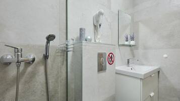 A modern bathroom with a glass shower enclosure, a handheld showerhead, a hairdryer, and a sink with a mirror above it.