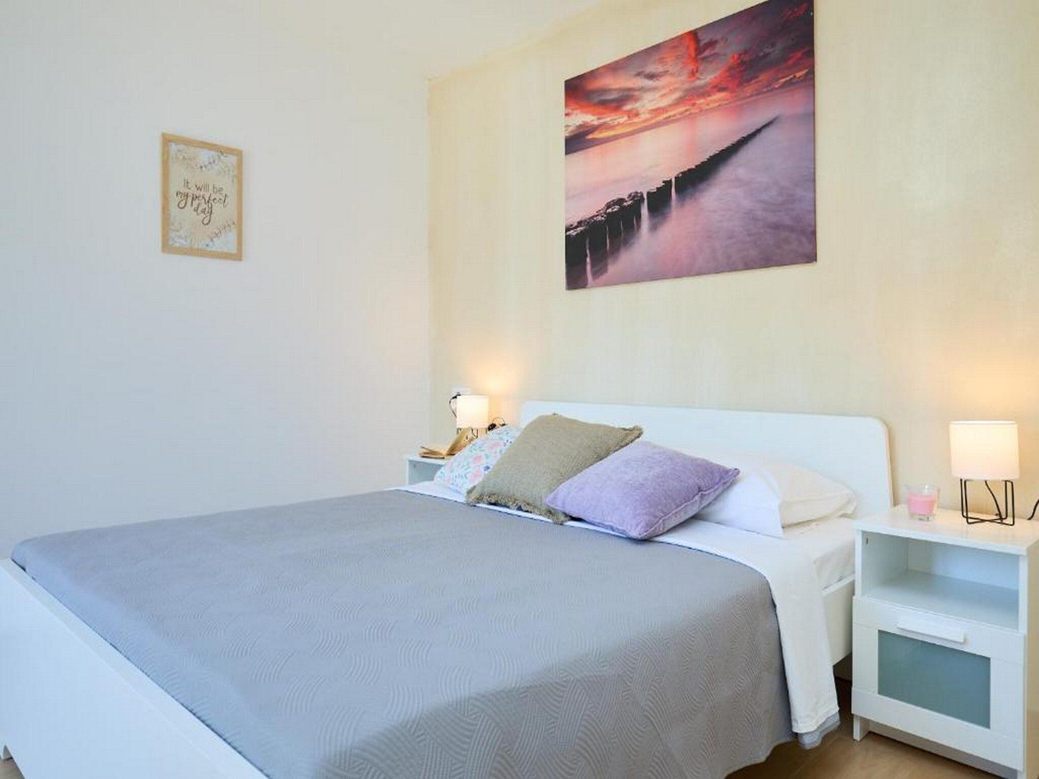 A neatly arranged bedroom with a large bed, two bedside tables with lamps, a framed quote on the wall, and a sunset painting above the bed.