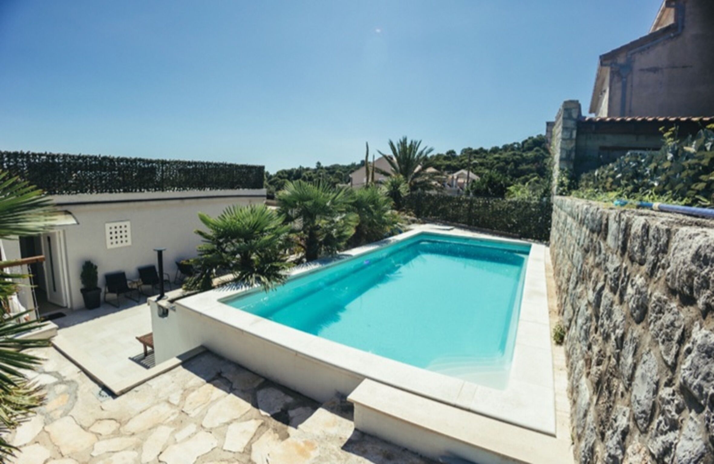 A rectangular swimming pool with a stone border, surrounded by a stone patio, and a stone wall on one side.