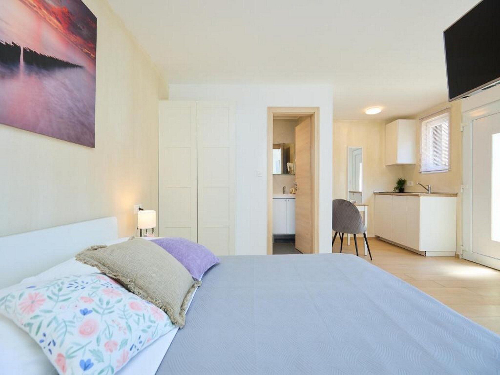 Een slaapkamer met een bed, nachtkastje en schilderij aan de muur. Er is een doorgang naar een keuken met een spoelbak en kastjes.