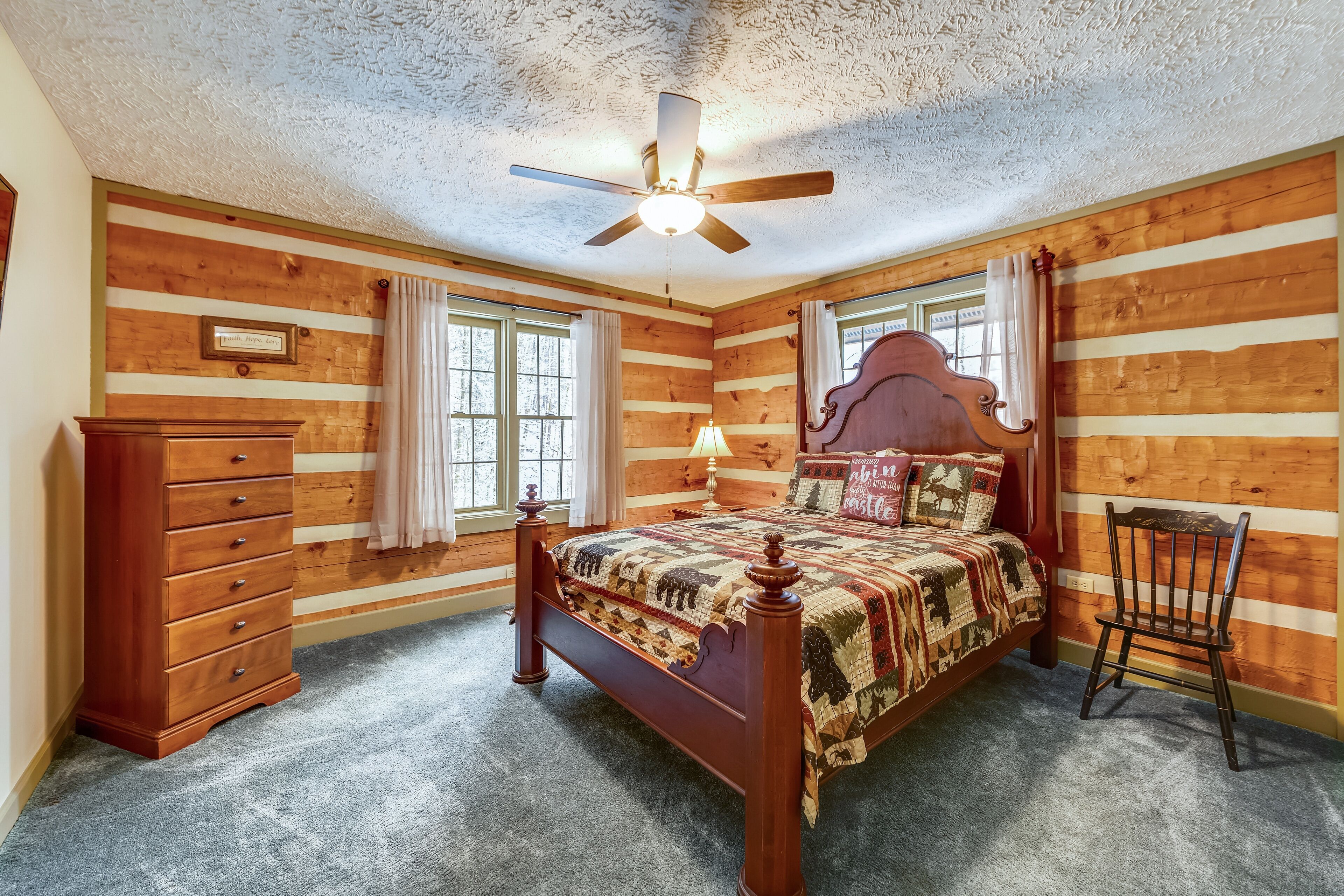 A log cabin-style bedroom with a wooden bed, dresser, and chair, featuring a ceiling fan and large windows.