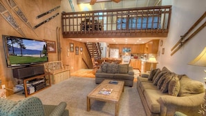 A cozy living room with a television, sofas, a coffee table, and a kitchen in the background.