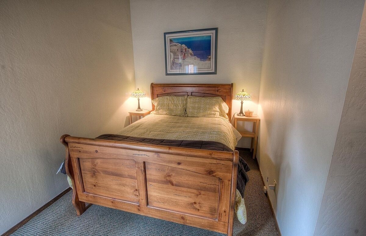 Uma cama de madeira com cabeceira, duas mesas de cabeceira com abajures e um quadro na parede.