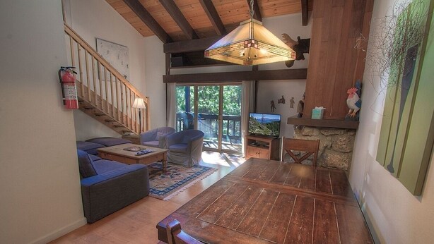 A spacious living room with a wooden dining table, a sofa, and a television. A staircase leads to an upper level, and a fire extinguisher is mounted on the wall. - Alpine Getaway 3 Bedroom Home by RedAwning (Tahoe City)