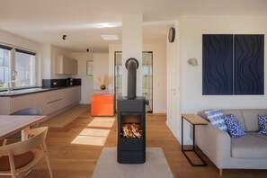 Une cuisine moderne dotée d’un poêle à bois central, un coin repas avec des chaises en bois et un espace de vie avec un canapé et des coussins.