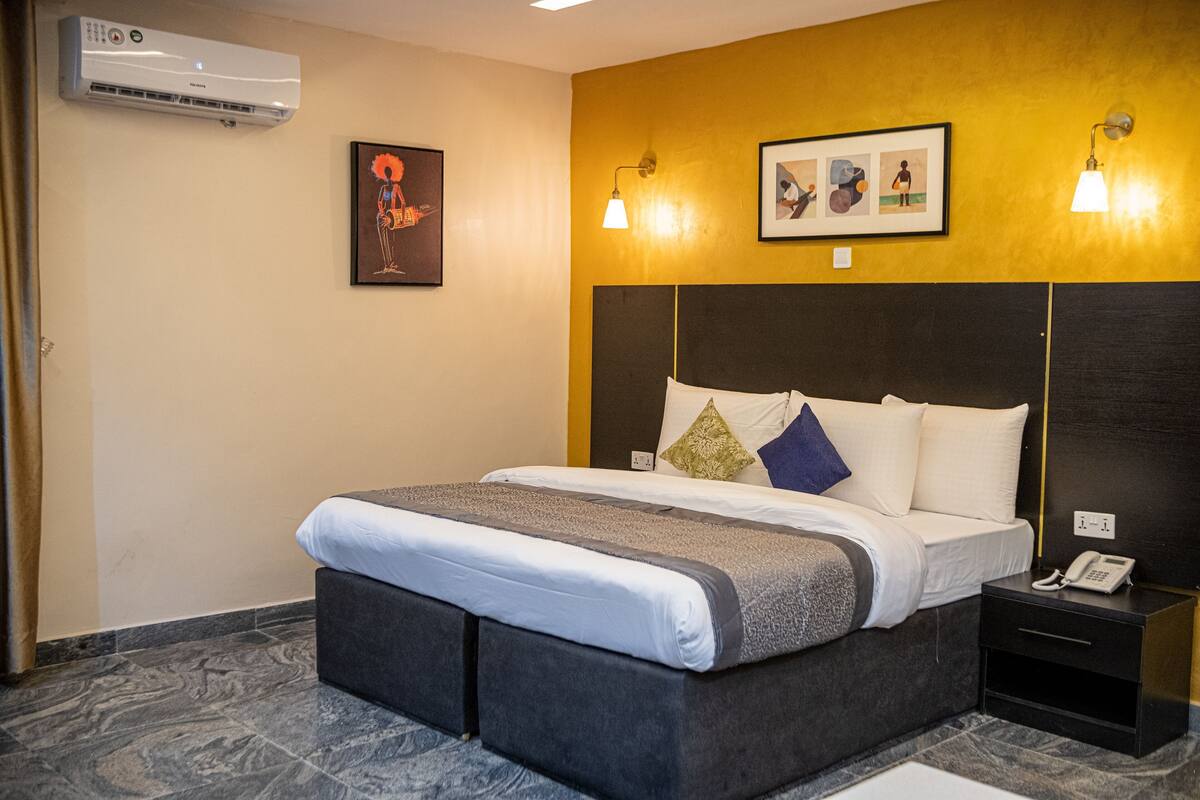 A hotel room with a bed, bedside table, wall-mounted air conditioner, and artwork.