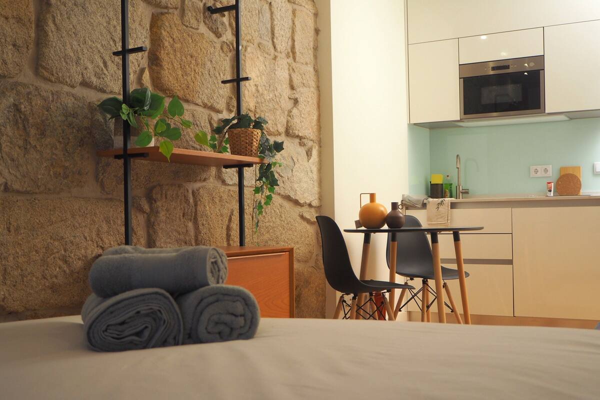 A modern room with a bed, a small table with chairs, a kitchen area with a microwave, and a stone wall.