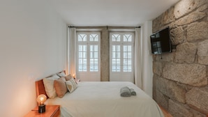 A bedroom with a large bed, a nightstand with a lamp, a stone wall, and a TV mounted on the wall. - Casa Farrapos (Porto)