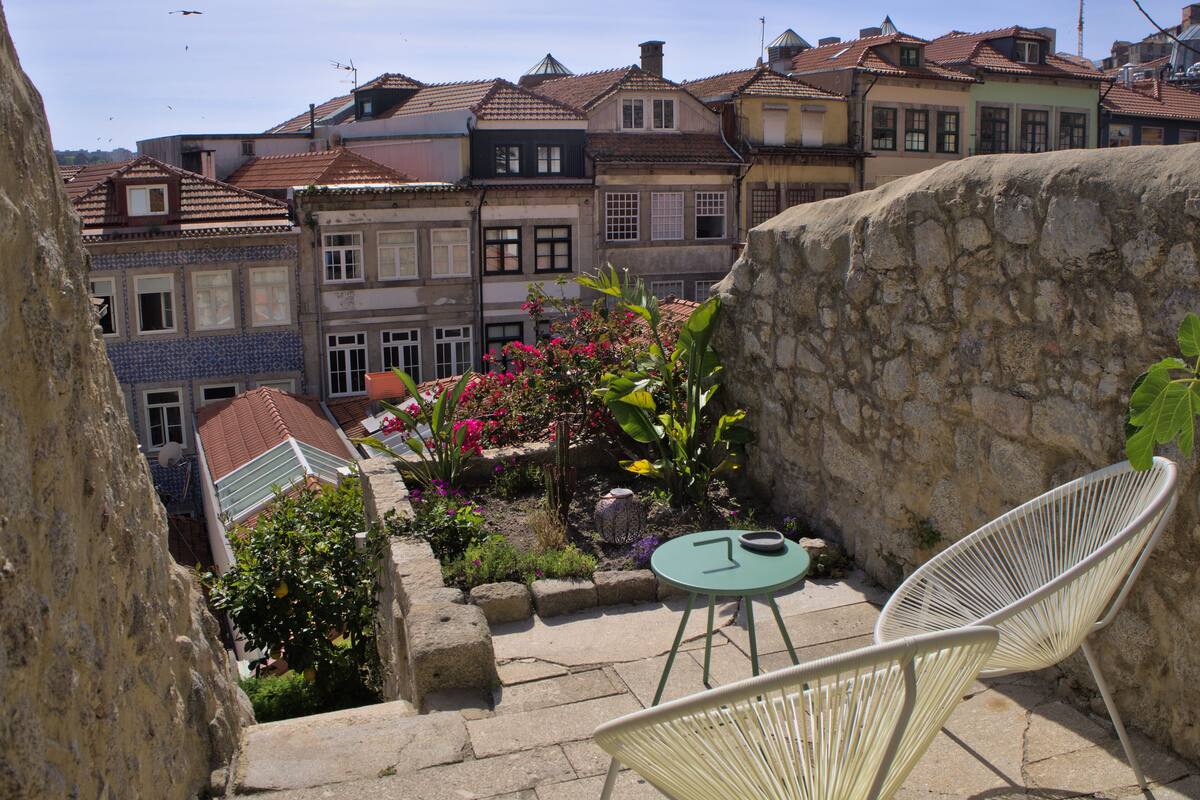 A courtyard with a stone wall, a small table, two chairs, and a variety of plants.