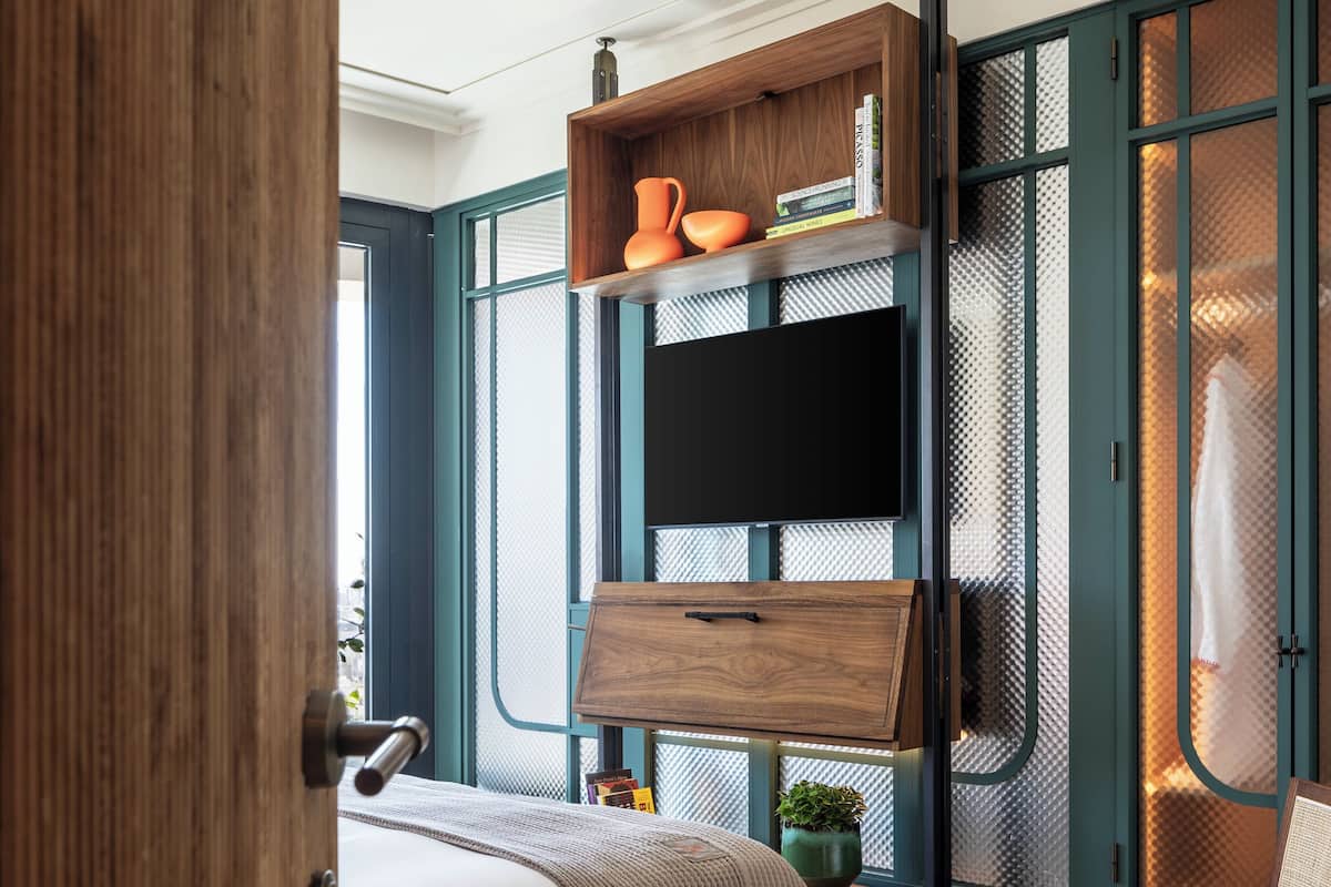 A modern bedroom with a bed, a TV mounted on a wall, a wooden shelf with books and a vase, and a chair.