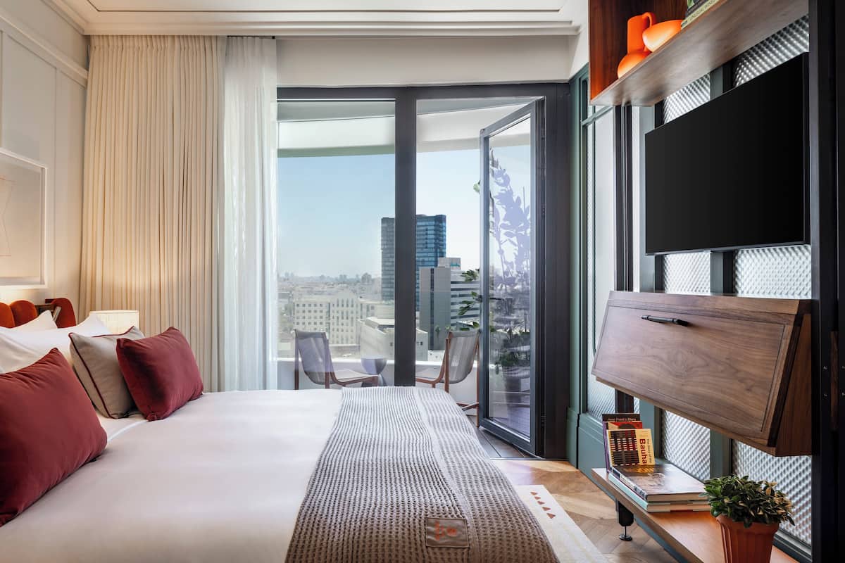 A modern hotel room with a large bed, a flat-screen TV, a sliding glass door to a balcony, and a city view.