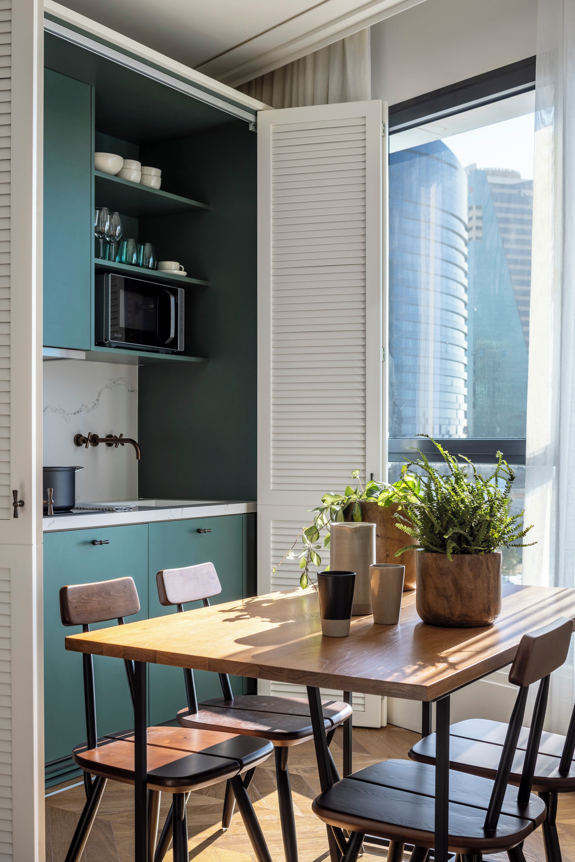 A modern kitchen with teal cabinets, a wooden dining table, and a view of tall buildings through a window.