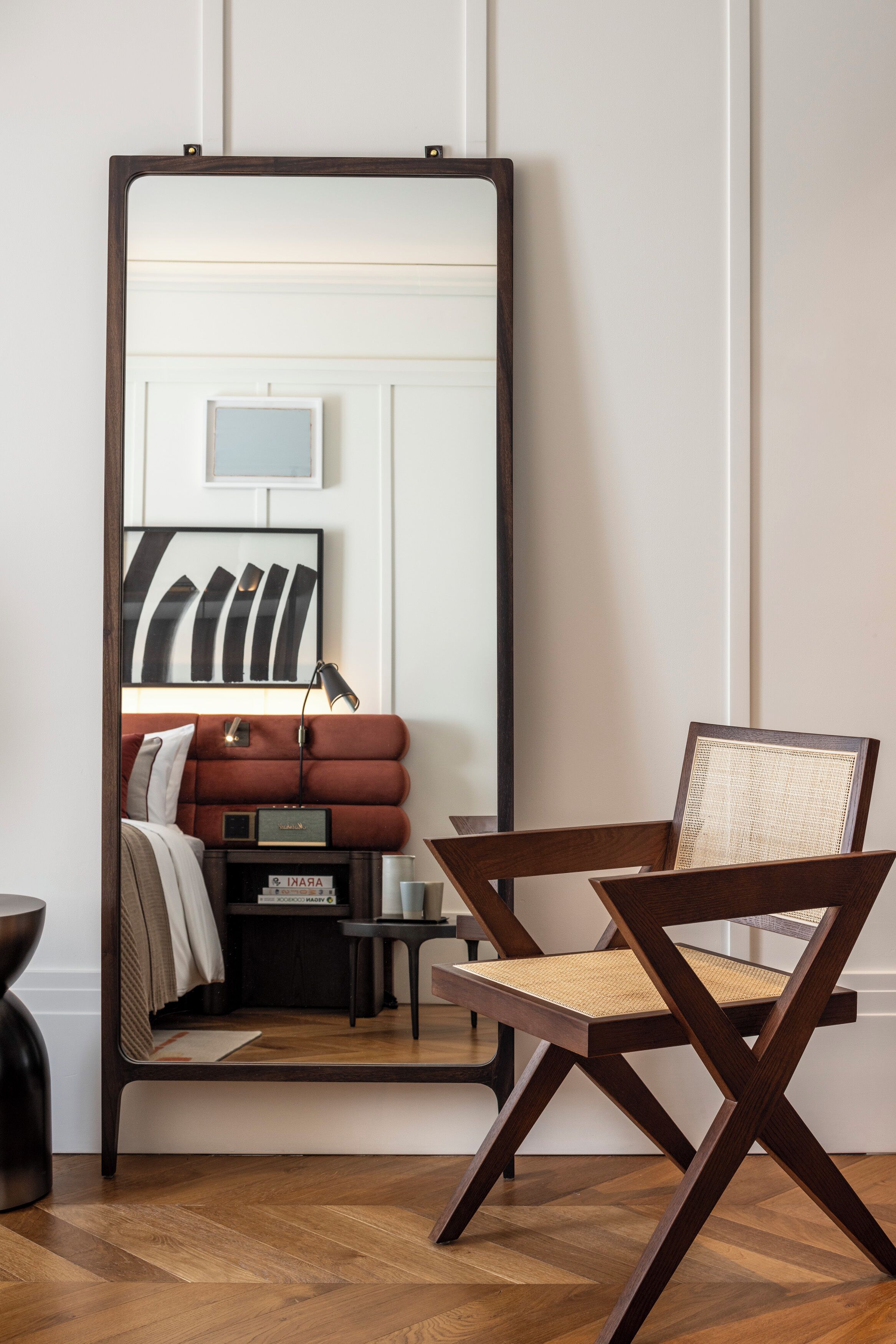 Una habitación con un espejo grande que refleja una cama, una silla y una mesita de noche con una lámpara.